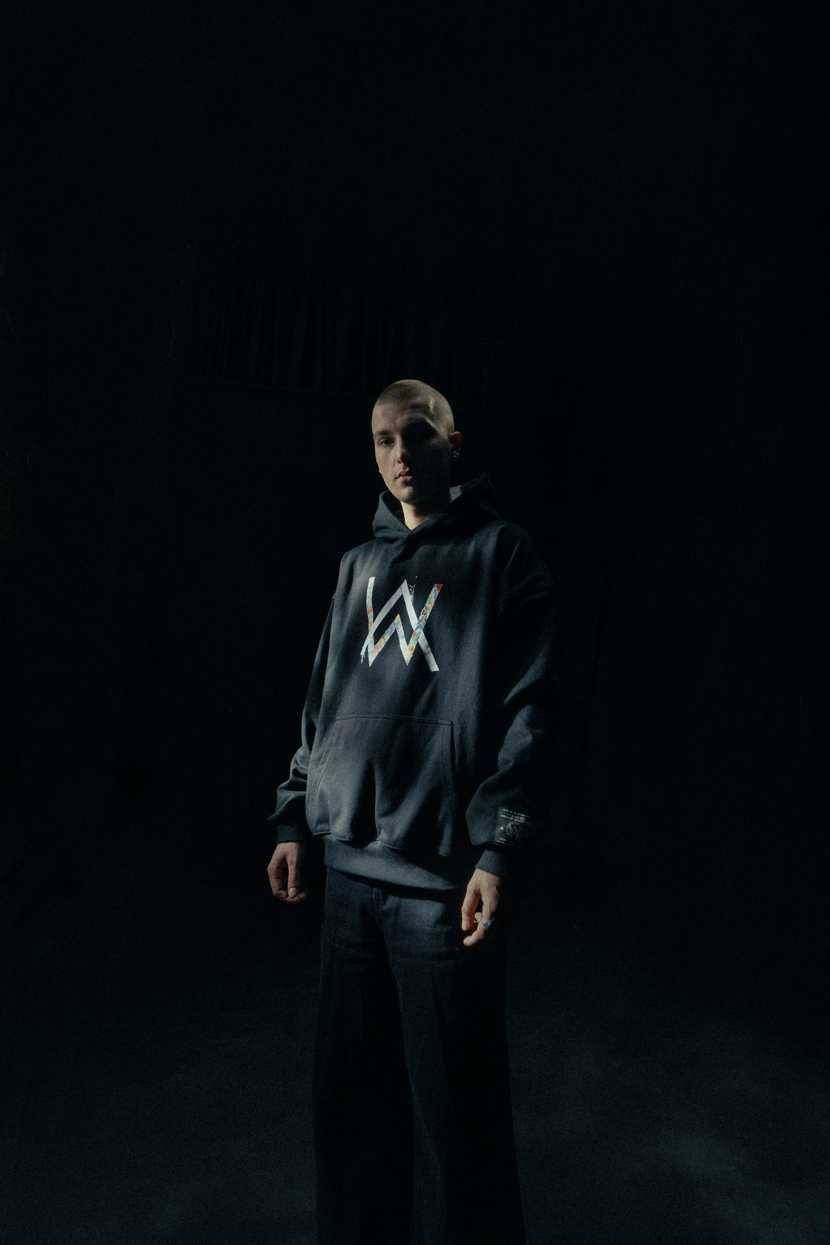 Alan Walker Tired 0 Hoodie worn by a model in a dark room, black hoodie with glowing colourful AW chest logo