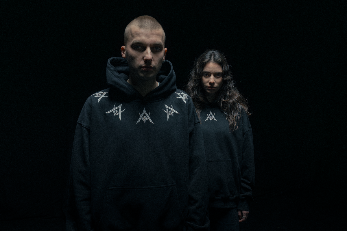 Alan Walker Watch The Drone Hoodie on two models in shadow, matching black hoodies with silver chest symbols