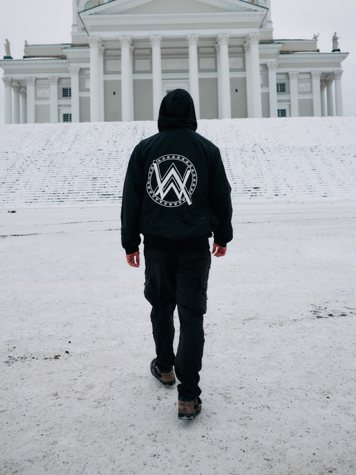 Alan Walker Core LV1 Windbreaker worn in snow outside a cathedral, white AW crest on the black hooded back
