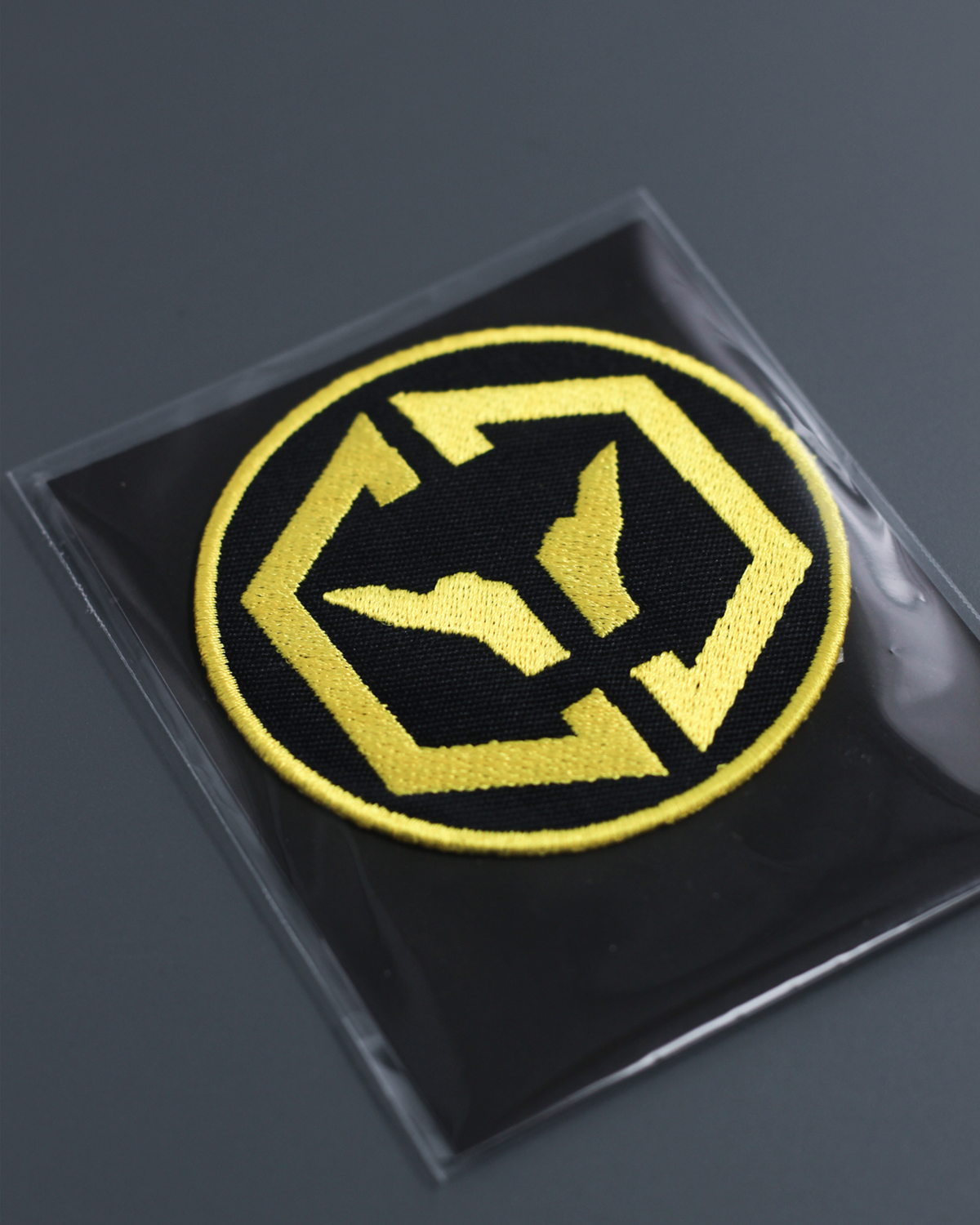 Alan Walker CheckPoint One Tribe Bundle patch shown at an angle, yellow embroidered crest in a clear pack