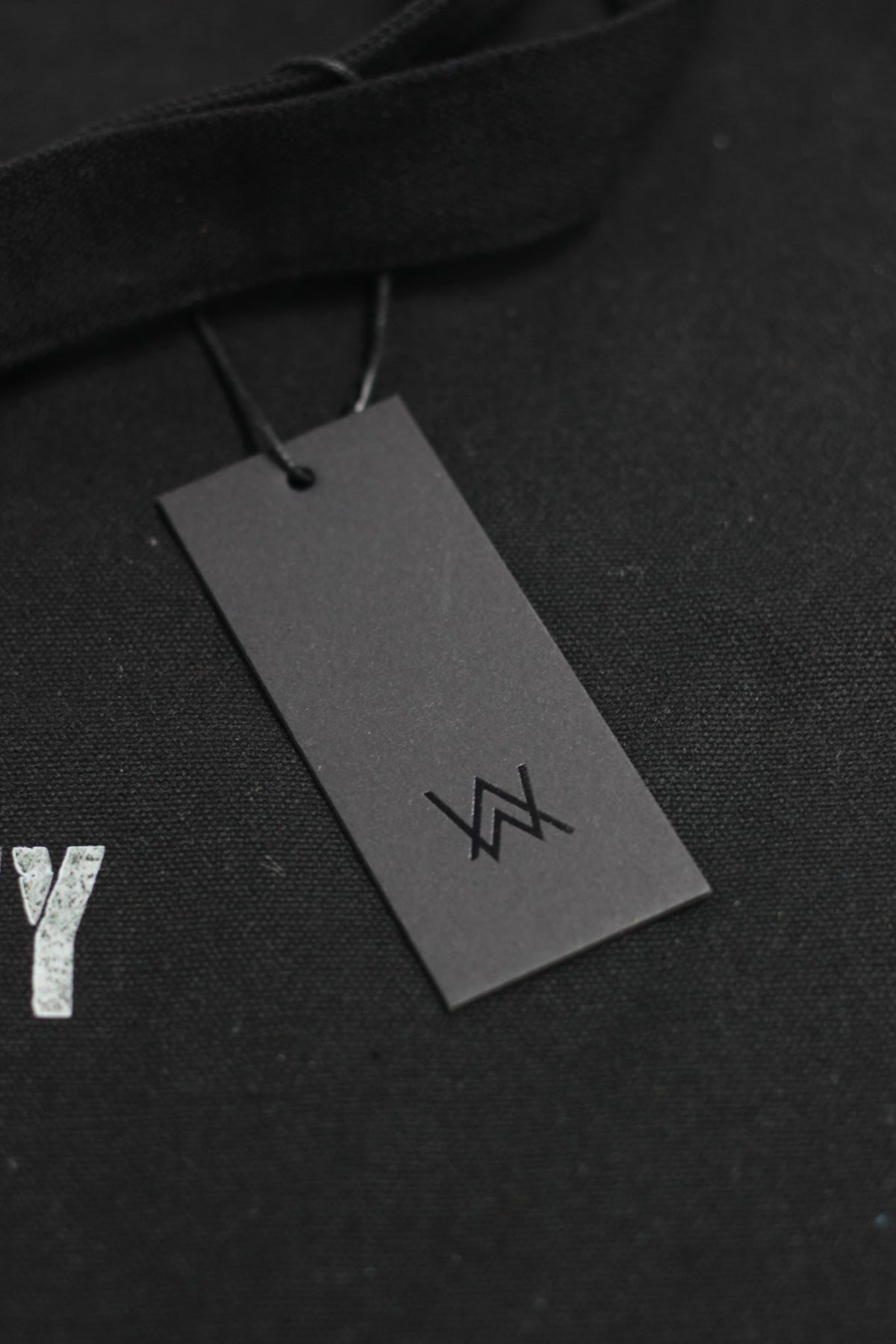 Alan Walker Tired Unity Tote hang tag detail, grey AW logo swing ticket resting on the black canvas bag