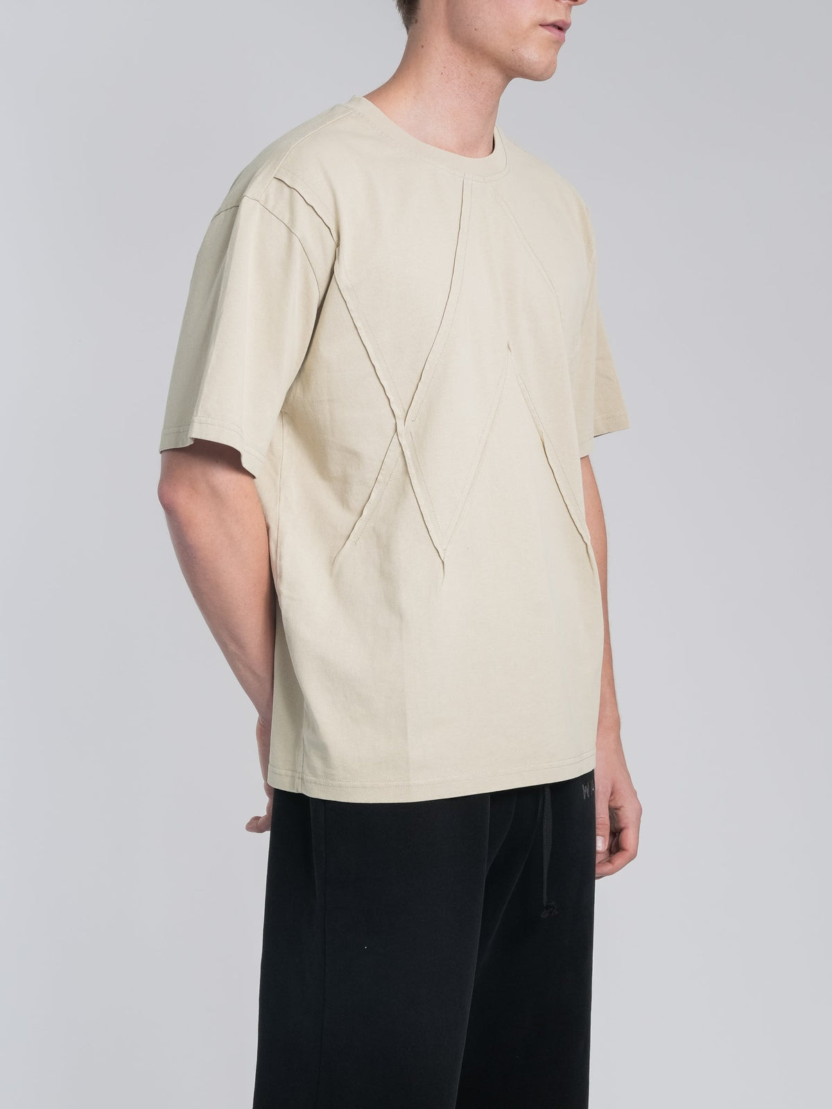 Alan Walker Stitch Logo Tee side view on a model, sand beige cotton tee showing the raised stitch panel lines