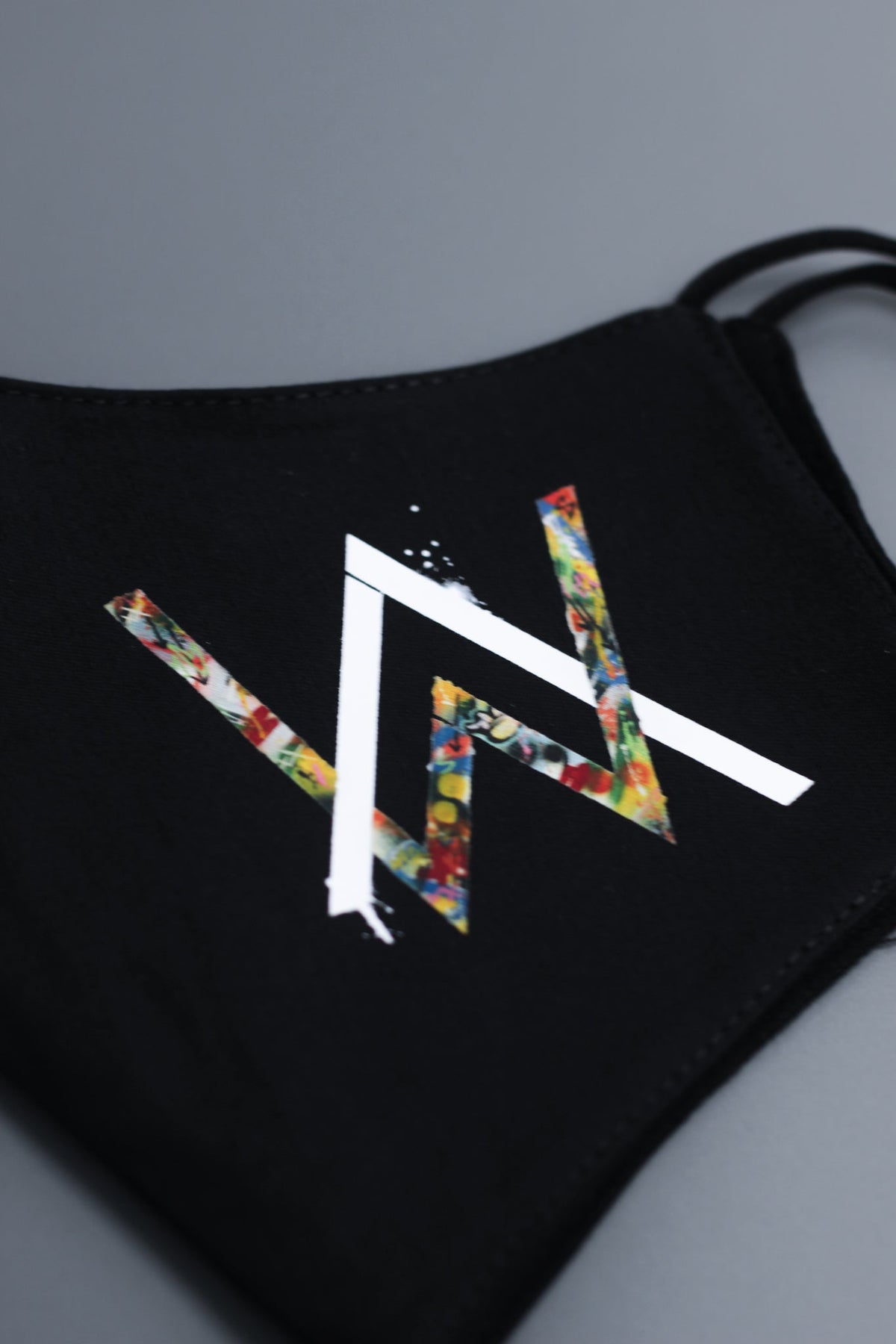 Alan Walker Tired Mask logo close-up, white AW emblem with multicoloured floral fill on black stretch fabric