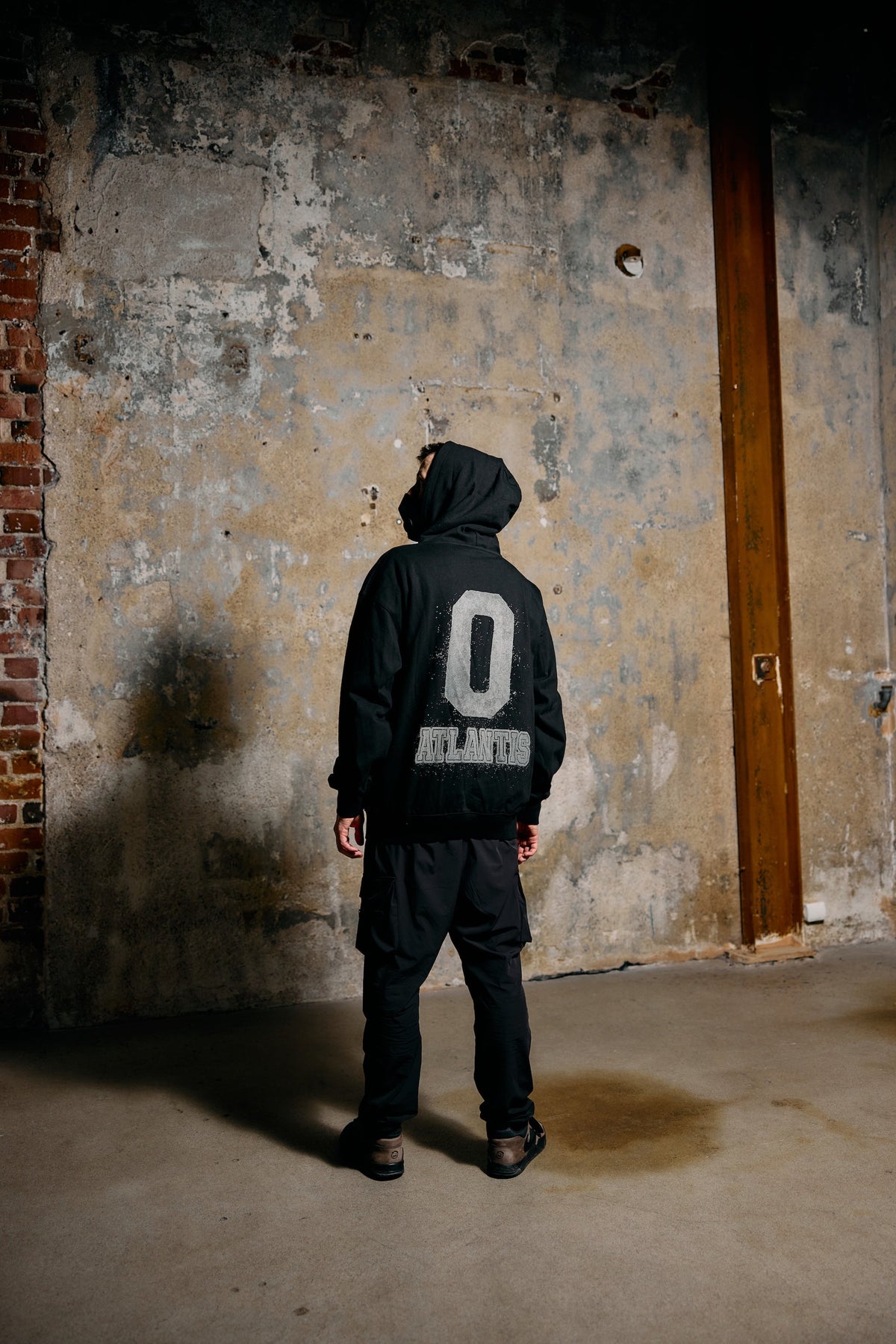 Alan Walker Atlantis 0 Hoodie lifestyle shot, fan in the black hood facing a weathered wall, 0 Atlantis back print