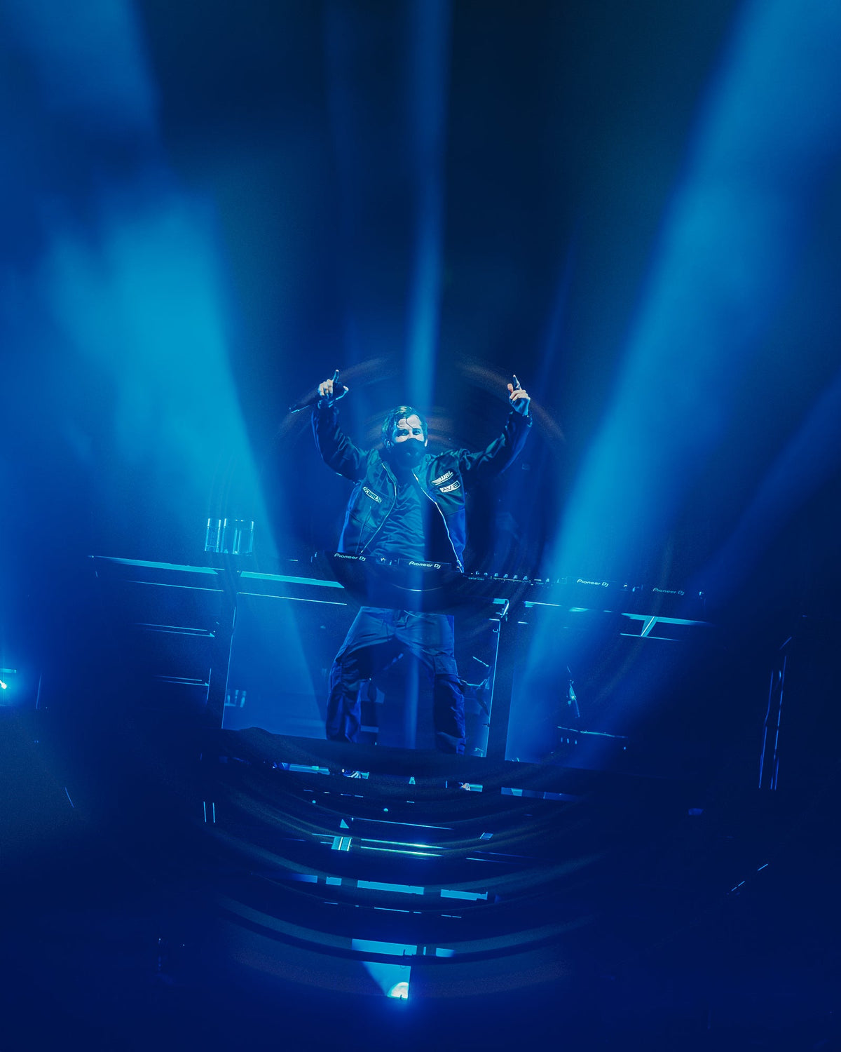 Alan Walker F1 Stage Jacket worn live under blue stage beams, masked DJ with arms outstretched in smoke