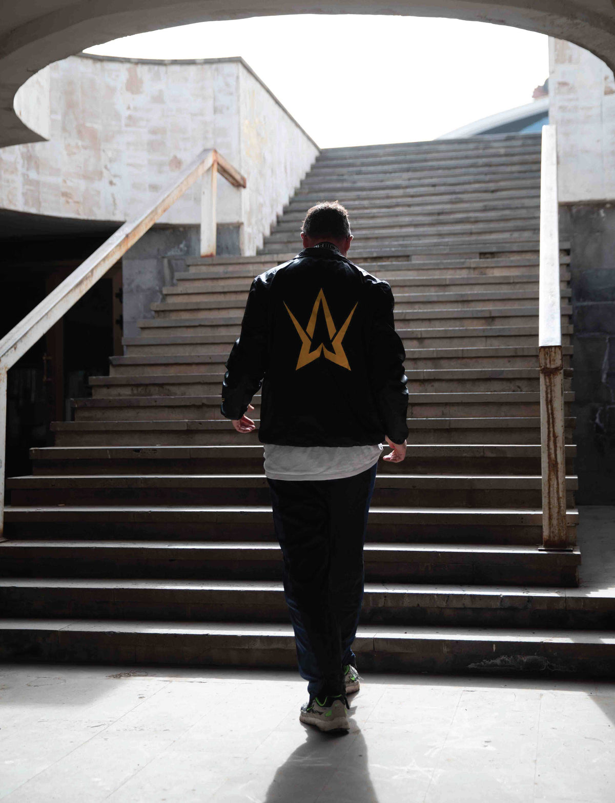Alan Walker Walkerverse Crew Jacket seen from behind on a model climbing stairs, gold Walker logo on black