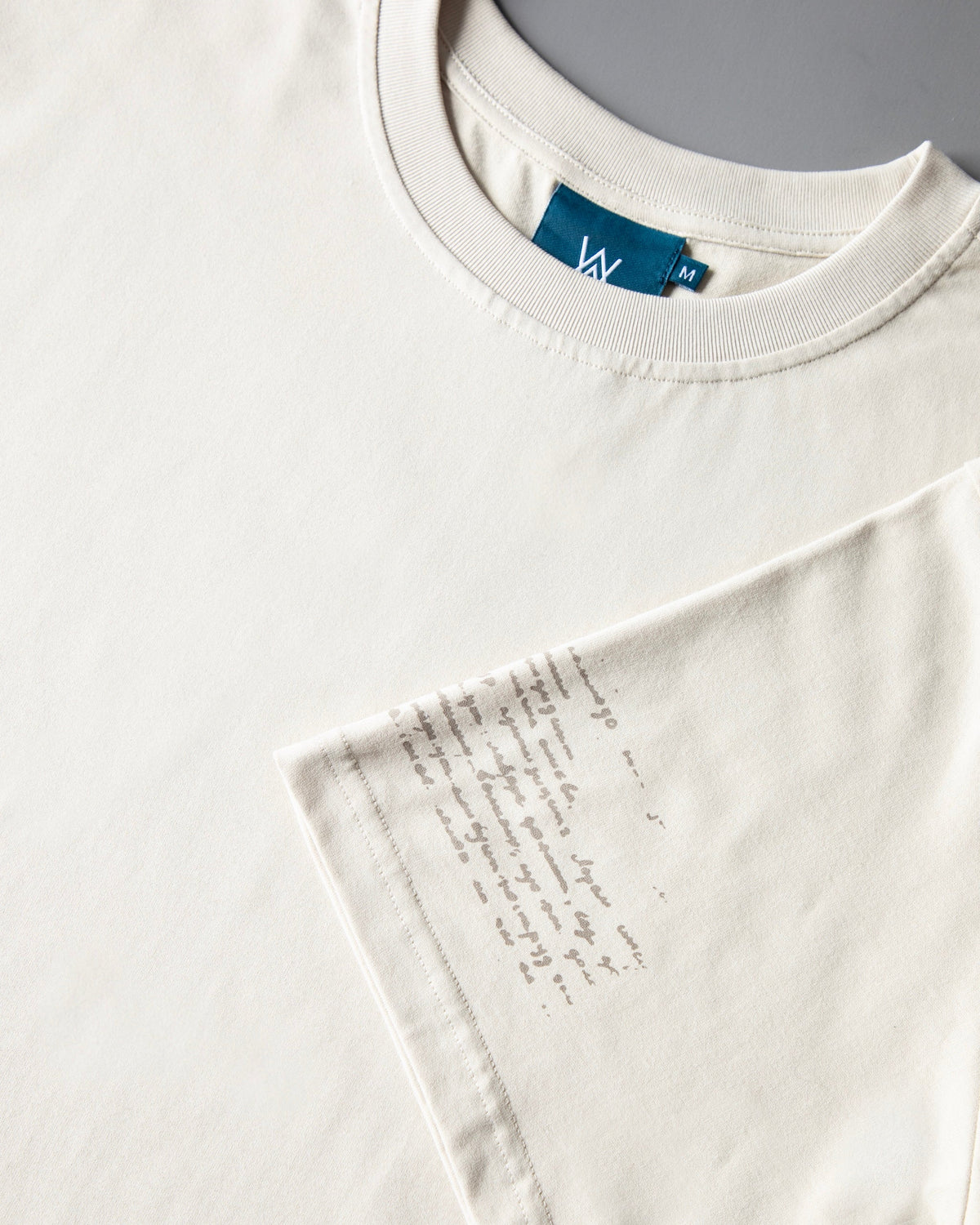 Alan Walker Encyclopedia T-Shirt folded, teal neck tag on off white cotton with faint handwritten sleeve text