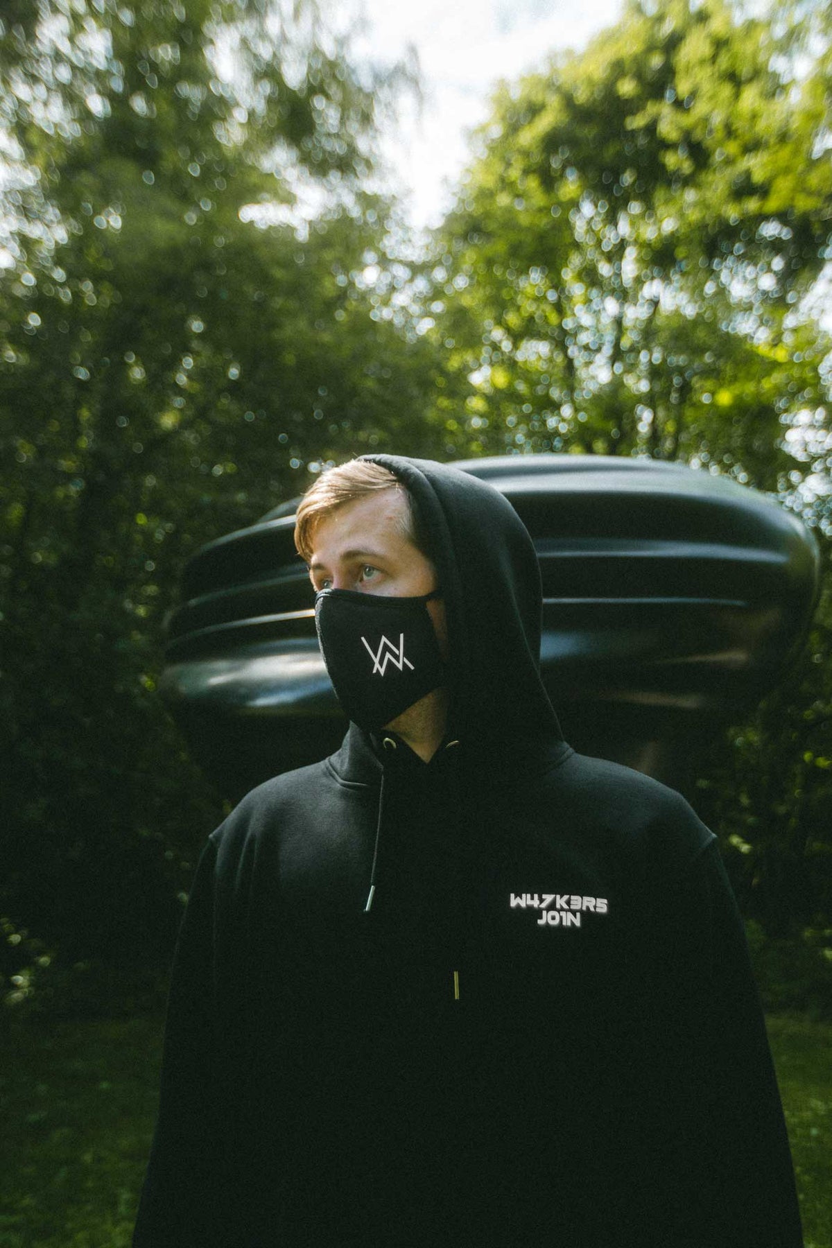 Alan Walker Core Logo Hoodie worn with the hood up and an AW face mask outdoors among green trees