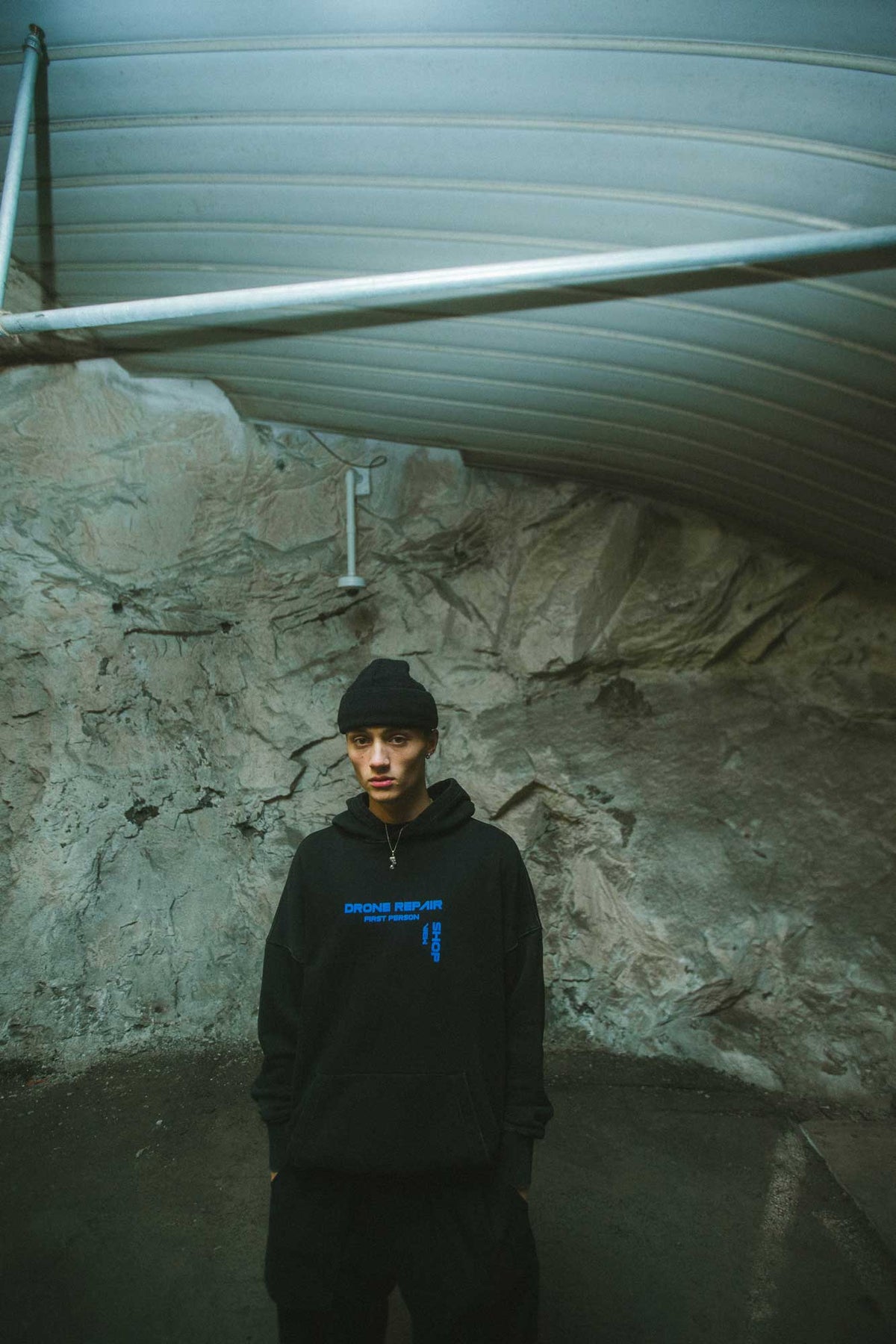 Alan Walker Drone Repair Shop Hoodie worn by a model in a rock tunnel, black hoodie with blue chest graphic
