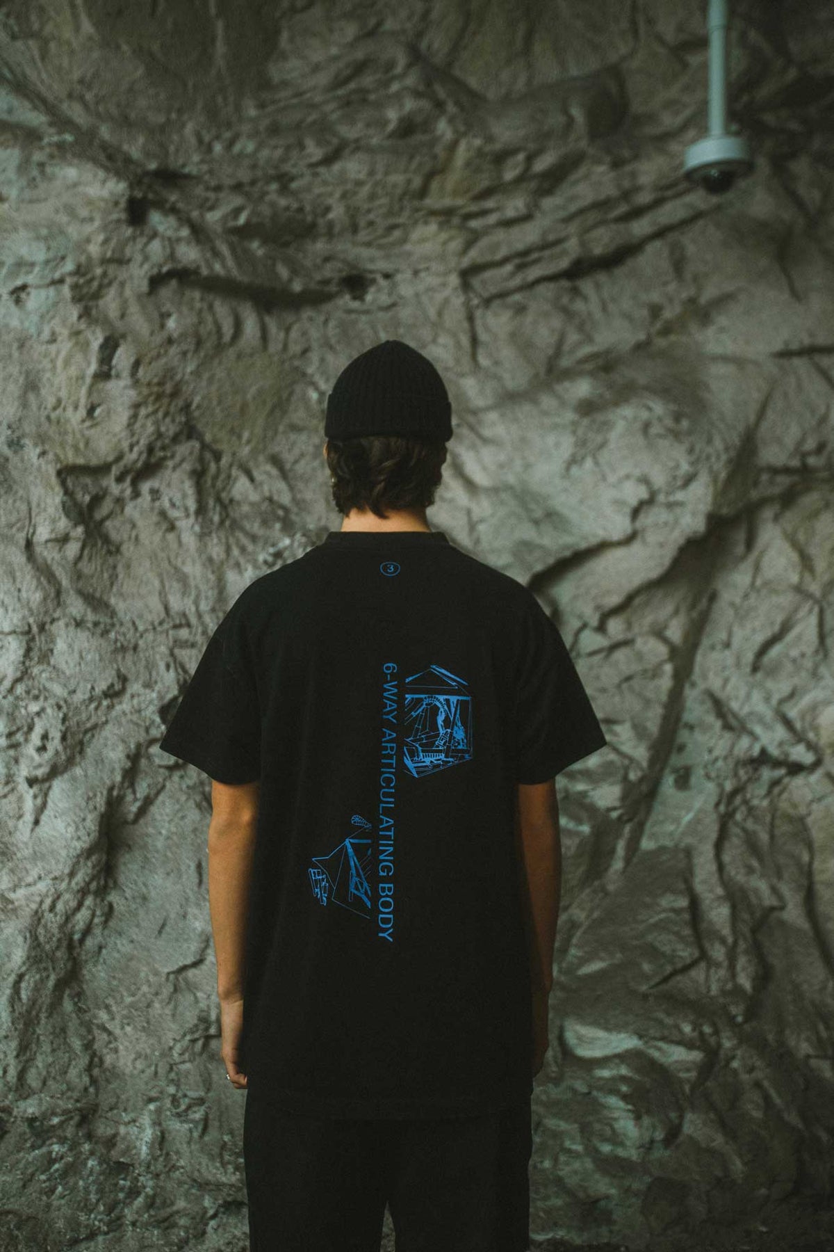 Alan Walker Drone Repair Shop T-Shirt back view on a model in a rock tunnel, blue drone graphic on black