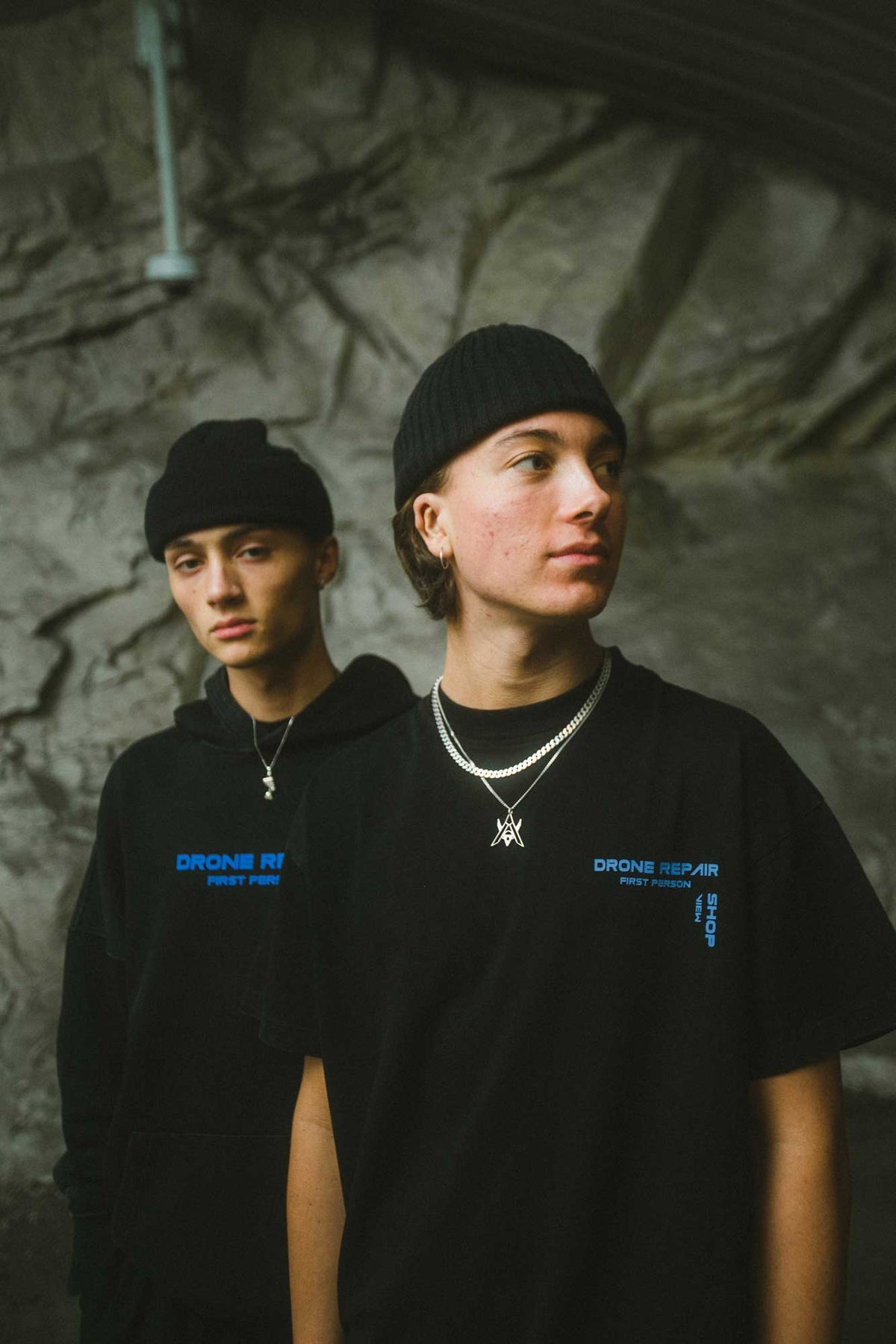 Alan Walker Drone Repair Shop T-Shirt worn by two models in a cave, blue chest prints on oversized black tees