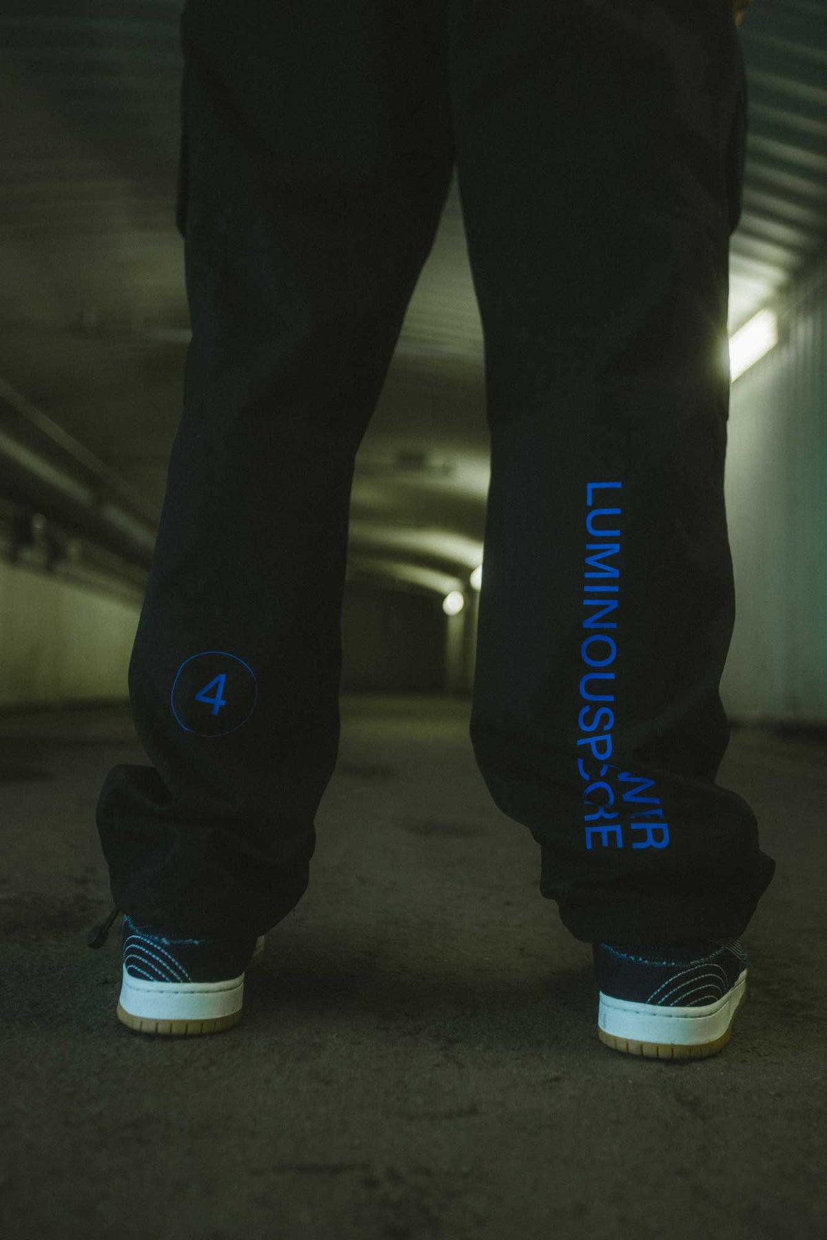 Alan Walker Drone Repair Shop Cargo Pants in black, worn in a lit tunnel with the blue Luminous Peace leg print