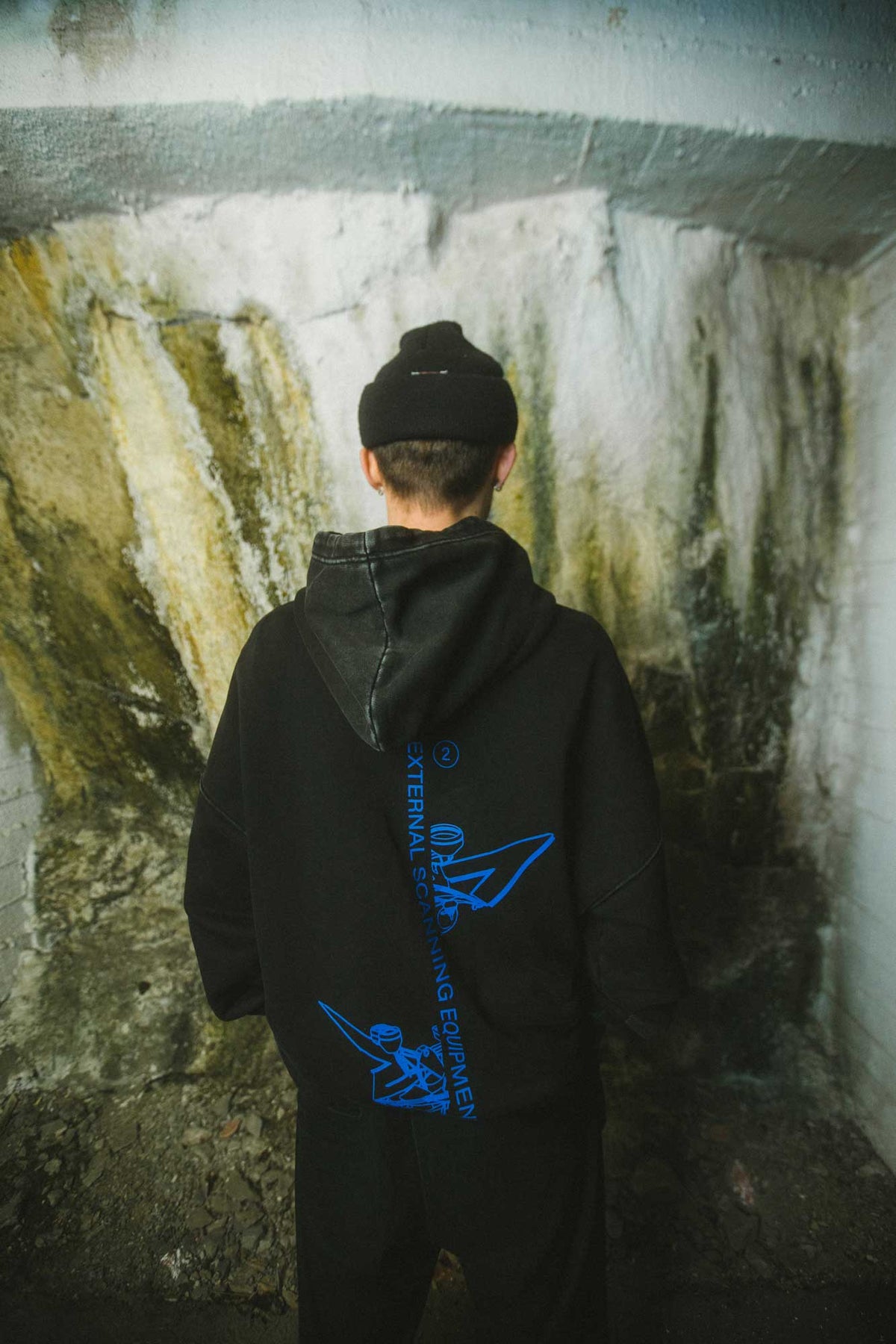 Alan Walker Drone Repair Shop Hoodie rear view on a model facing a cave wall, blue drone print down the back