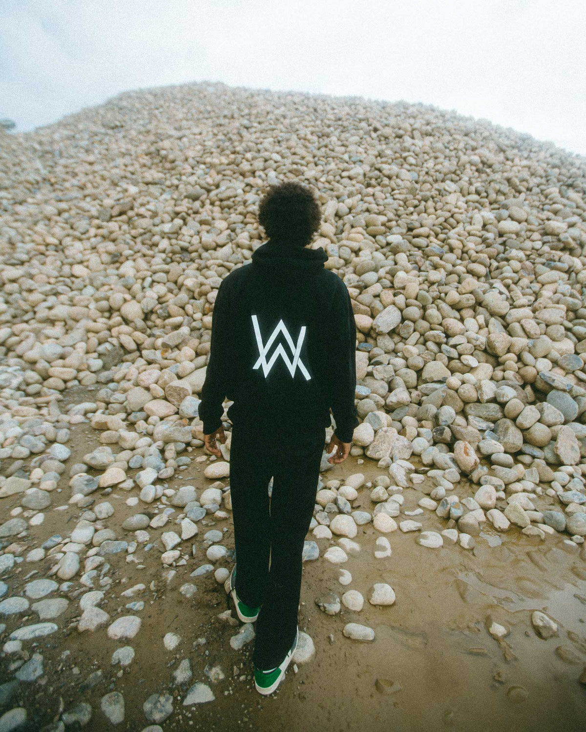 Alan Walker Core Logo Zip Hoodie worn on a pebble beach, back view with the white AW logo between shoulders