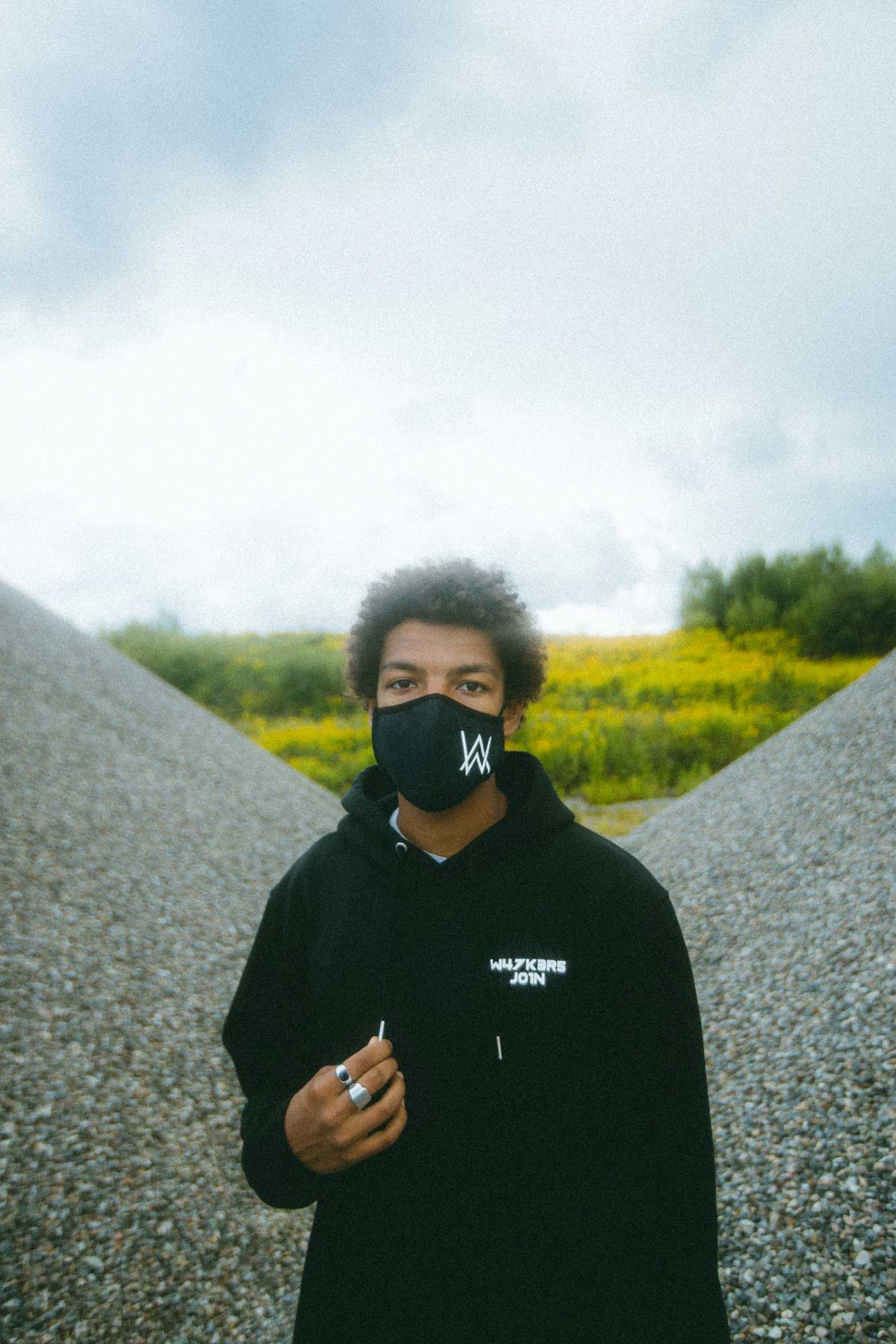 Alan Walker Core Logo Hoodie worn by a model on a gravel path, black pullover with chest text and AW mask