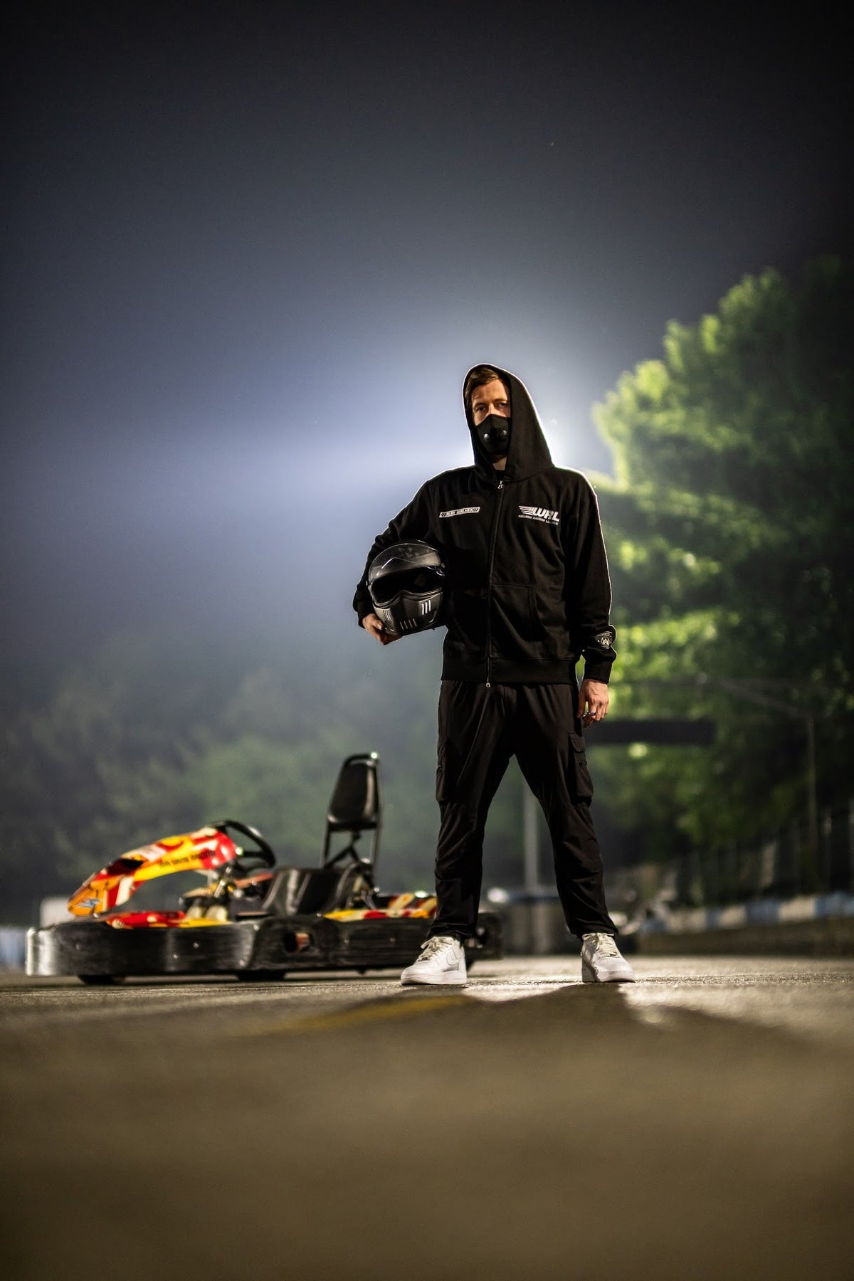 Alan Walker WRL Team Zip Hoodie on a model holding a helmet beside a go-kart at night, full black racing fit