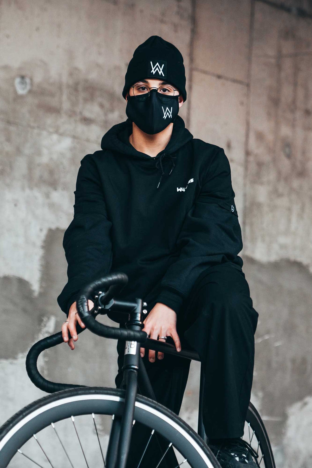 Alan Walker Core Logo Face Mask worn with a beanie by a cyclist at a concrete wall, AW logo in white