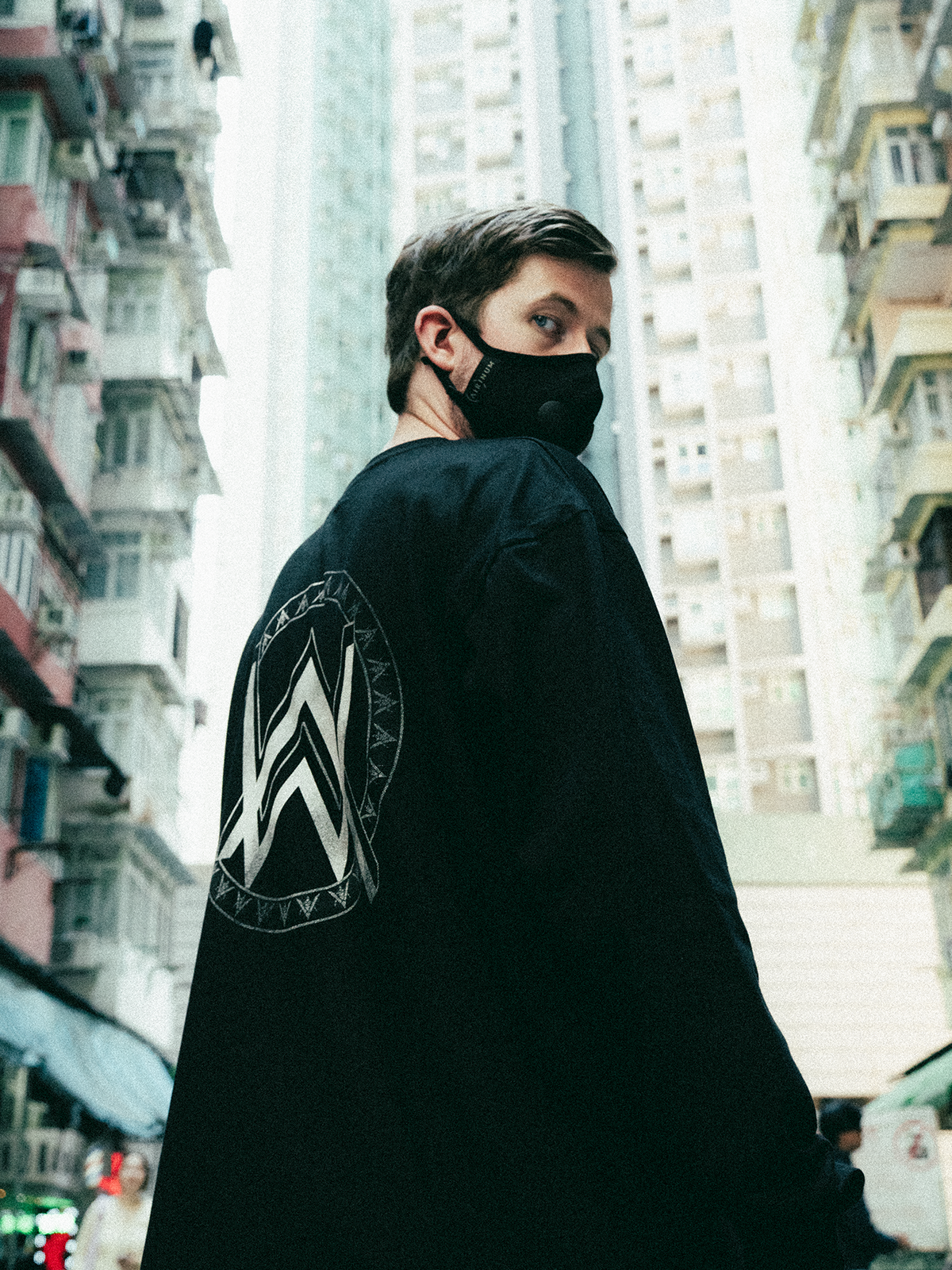 Alan Walker Monster Core Longsleeve worn by a masked model turning to show the AW crest, city high rise backdrop